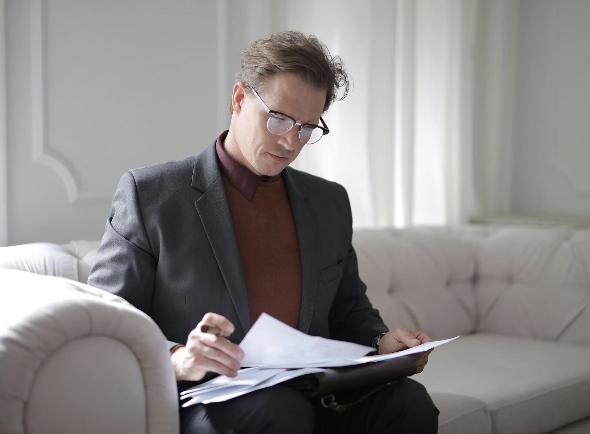 A professional man with glasses, wearing a dark blazer and turtleneck, focused on reviewing papers in his hand in a well-lit room.