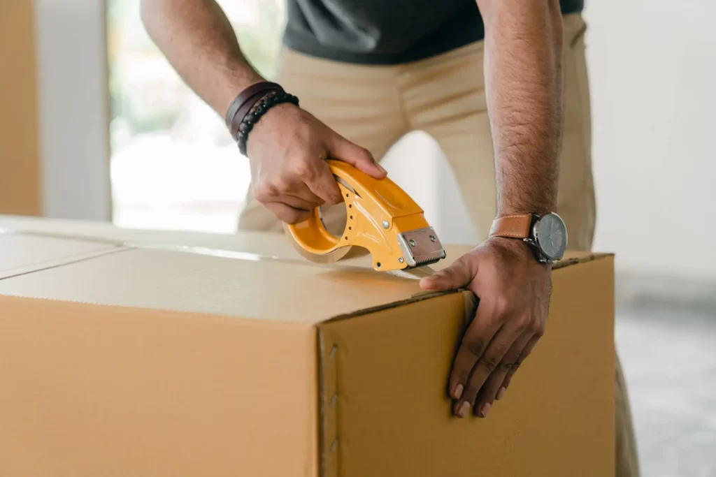 Close-up of a man's hands using a yellow tape dispenser to seal a cardboard box, highlighting the importance of secure packaging.