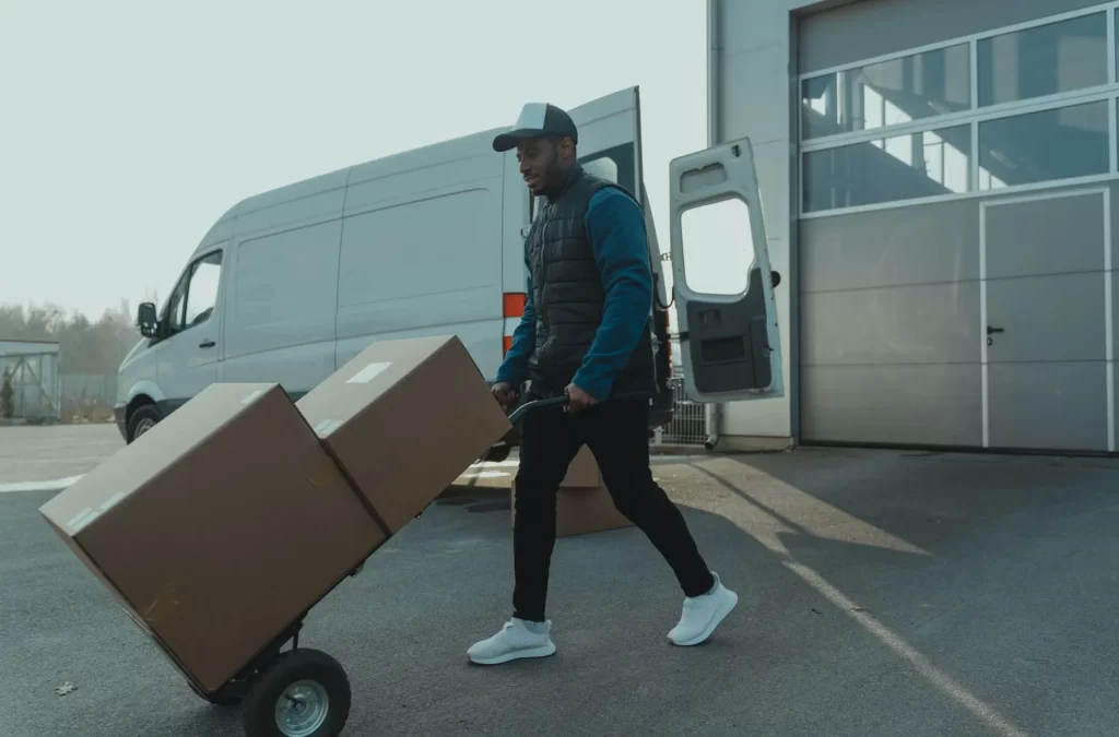 Man in a blue jacket and cap transporting a cardboard box on a dolly, highlighting the benefits of outsourcing trucking services for your business with a white van ready in the background.