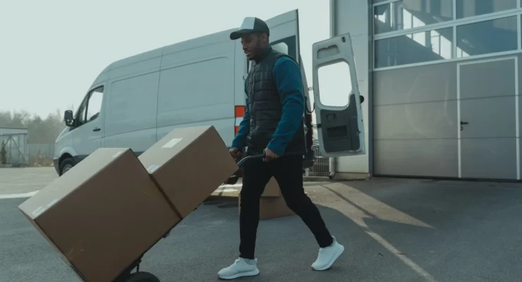 Man in a blue jacket and cap transporting a cardboard box on a dolly, highlighting the benefits of outsourcing trucking services for your business with a white van ready in the background.