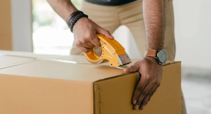 Close-up of a man's hands using a yellow tape dispenser to seal a cardboard box, highlighting the importance of secure packaging.